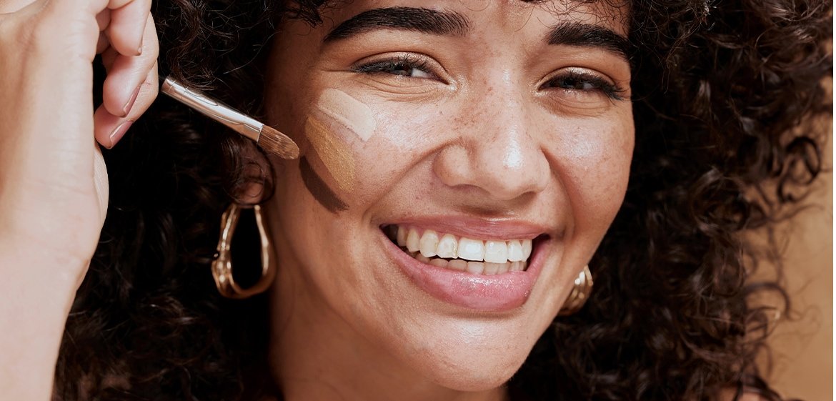 Junge Frau lacht und trägt Concealer in drei verschiedenen Farbtönen auf ihrer Wange.